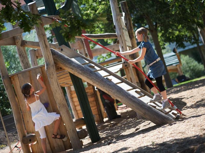 Kinder spielen auf einem hölzernen Klettergerüst mit Rutsche im sonnigen Garten.