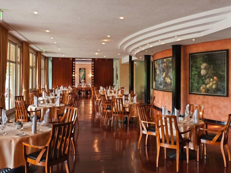 Elegantes Restaurant mit Holzboden, runden Tischen, weißen Tischdecken und großen Fenstern.