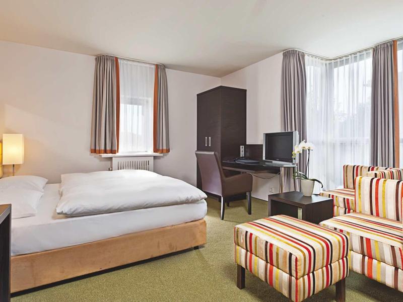 Modernes Hotelzimmer mit großem Bett, Schreibtisch, Fernseher und gestreiftem Sessel mit Hocker.