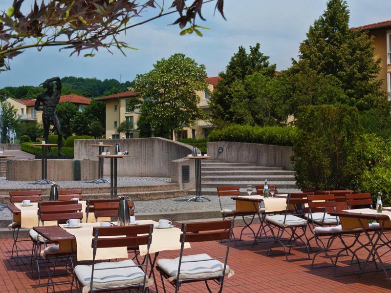 Leerer Außenbereich mit Stühlen und Tischen, Bäumen und Gebäude im Hintergrund bei Tageslicht.