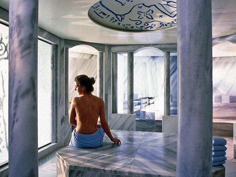 Frau sitzt in hellem, modernem Marmorraum mit Säulen und großem Fenster, trägt Handtuch um Hüfte.