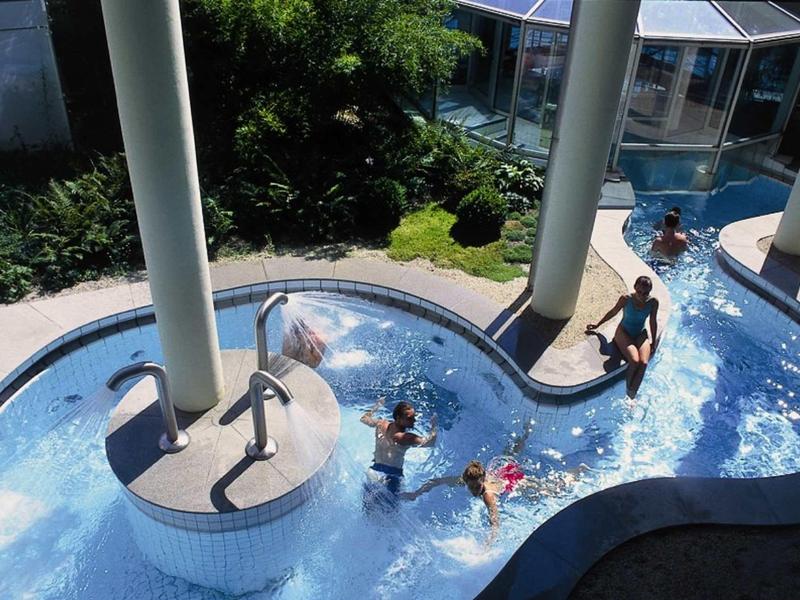 Wasserrutsche mit drei Kindern, die im klaren Wasser unter grauen Säulen rutschen, umgeben von Pflanzen.