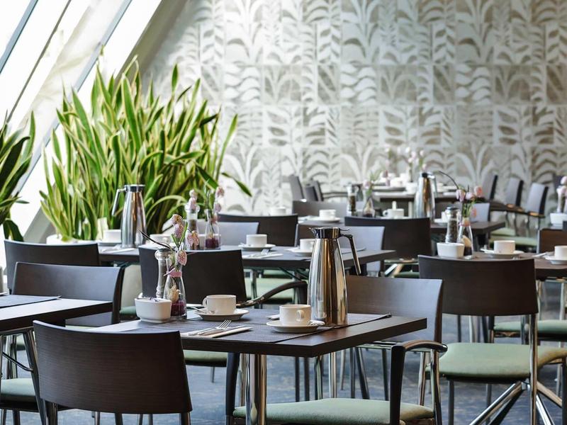 Modernes Restaurant mit grünen Pflanzen, dunkelbraunen Tischen und Stühlen vor gemusterten Wänden.