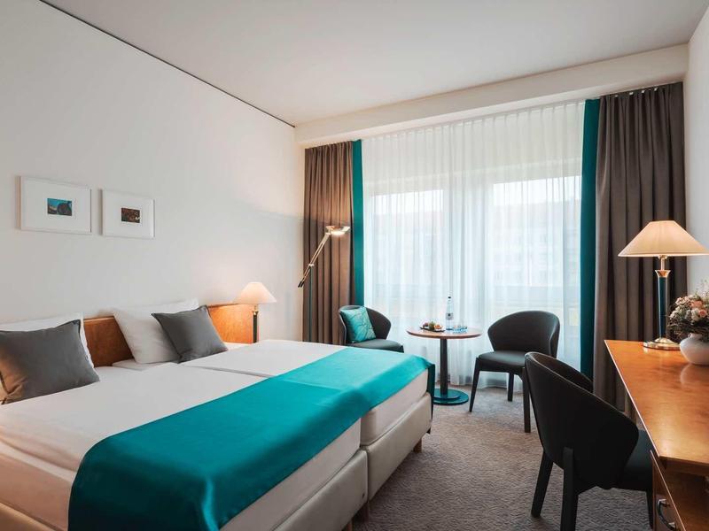 Modernes Hotelzimmer mit großem Bett, Holzschreibtisch, zwei Sesseln und bodentiefen Fenstern.