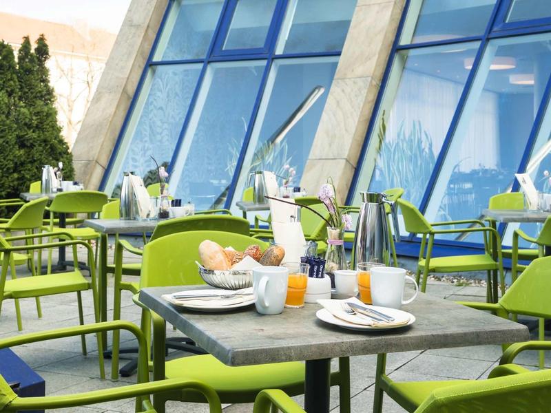 Outdoor-Café mit grünen Stühlen, Tischen, Frühstücksgeschirr und großen Glasfenstern im Hintergrund.