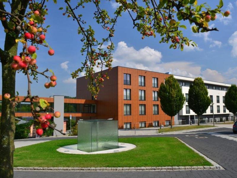 Moderne Gebäude mit roten und weißen Fassaden, Apfelbaum im Vordergrund, blauer Himmel mit Wolken.