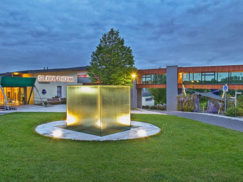 Moderner Park mit beleuchtetem Würfelbrunnen, grüner Wiese und Gebäuden bei bewölktem Himmel.