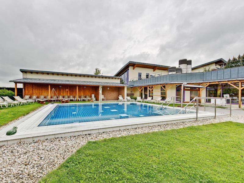 Moderner Pool mit Terrasse vor großen Holzhäusern unter bewölktem Himmel.