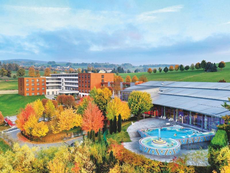 Bunte Herbstbäume umgeben ein großes Gebäude und einen Außenpool mit Wasserspiel vor grünen Hügeln.