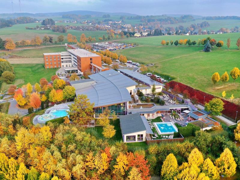 Landschaft mit buntem Herbstwald, mehreren Gebäuden, Schwimmbädern und grünen Wiesen im Hintergrund.