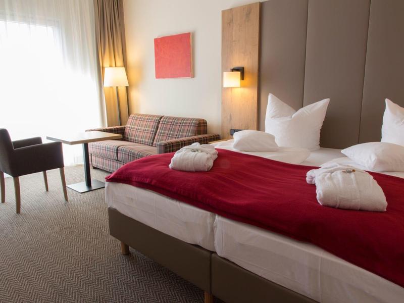 Gemütliches Hotelzimmer mit zwei Betten, roten Decken, Sofa, Tisch, Stühlen und großen Fenstern.