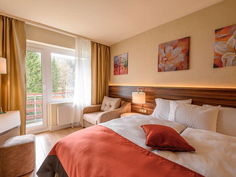 Gemütliches Schlafzimmer mit Doppelbett, oranger Bettdecke, Sesseln und Balkon mit natürlichem Licht.