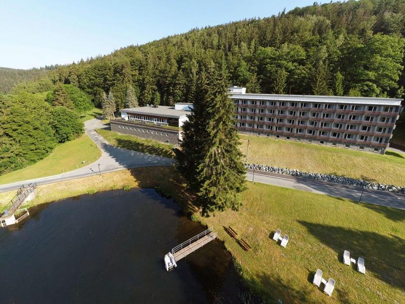 Modernes Hotel am Waldrand mit See, Steg und Liegestühlen auf einer Wiese.