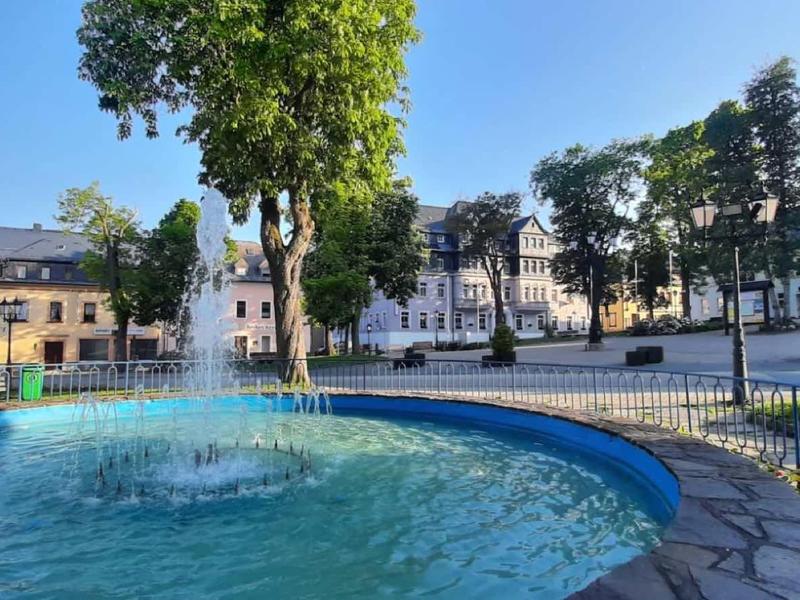 Runder blauer Springbrunnen mit Wassersäule, umgeben von Bäumen und weißen Gebäuden bei klarem Himmel.