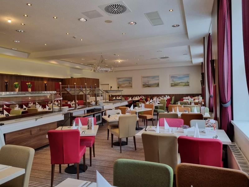 Hell eingerichteter Speisesaal mit roten und beigen Stühlen und langen Fenstern mit Vorhängen.