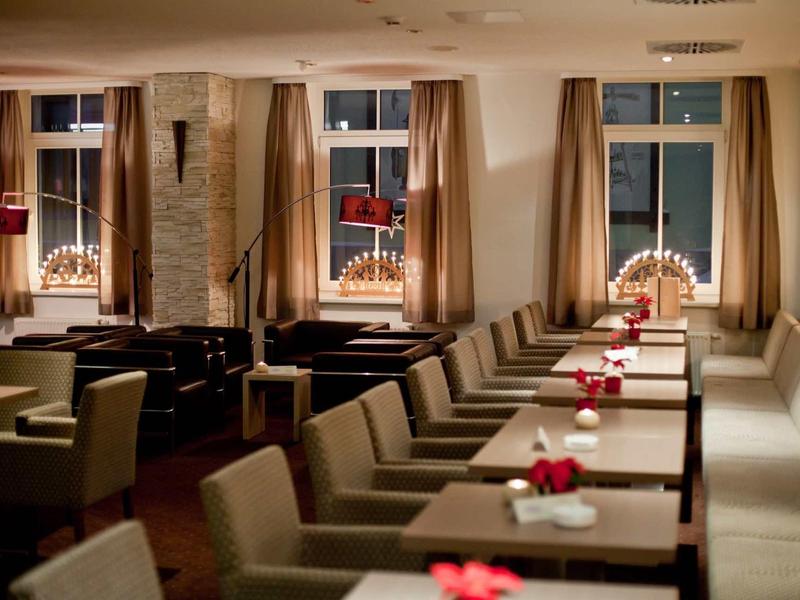 Gemütlicher Gastraum mit braunen Sesseln, langen Tischen, roten Tischdekorationen und Fensterbeleuchtung.