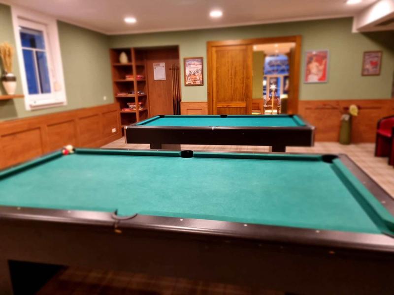 Ein Raum mit zwei grünen Billardtischen, Holzwänden und Regalen, beleuchtet durch Deckenlampen.