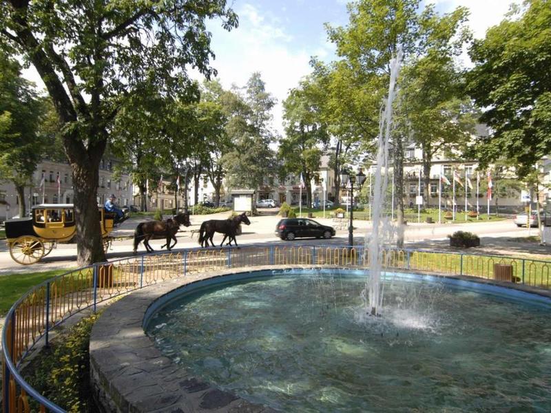 Runder Brunnen mit spritzendem Wasser, umgeben von Bäumen und Pferdekutsche auf Straße dahinter.