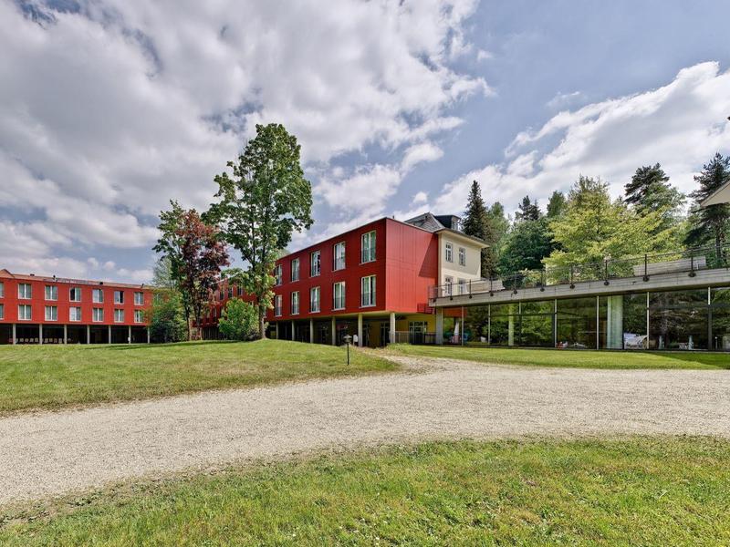 Modern hotel with red buildings surrounded by green grass and trees under a cloudy sky.