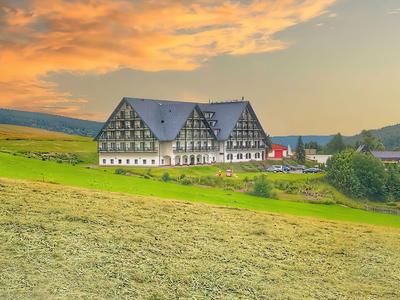 Großes Hotelgebäude mit Fachwerk vor einer grünen Landschaft und einem orangefarbenen Sonnenuntergang.