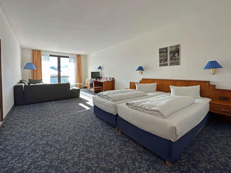 Spacious hotel room with two single beds, sofa, desk, and blue carpet floor.