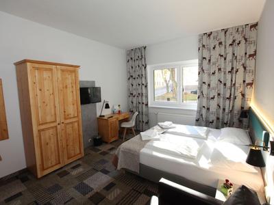 Helles Hotelzimmer mit Doppelbett, Holzschrank, Schreibtisch und Fenster mit gemusterten Vorhängen.