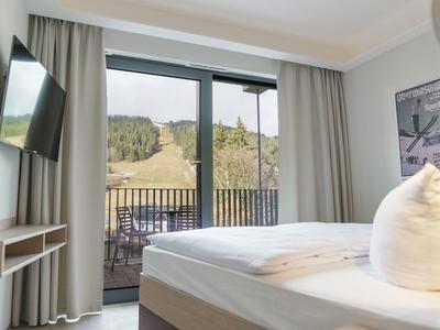 Hell eingerichtetes Hotelzimmer mit Balkon und Blick auf bewaldete Hügel.