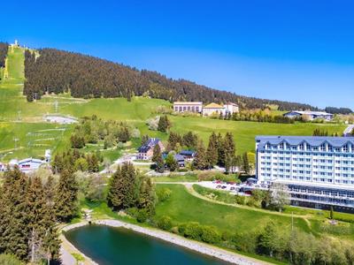 Hotel complex with lake in front of a forested hill under clear blue sky.