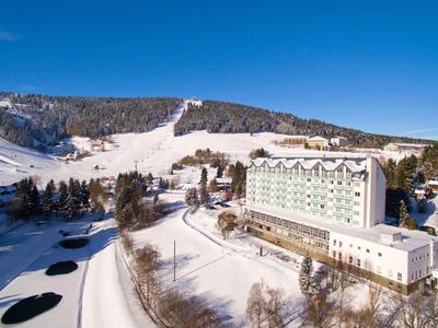 Large hotel beside snow-covered slopes and ski trails under clear blue sky.