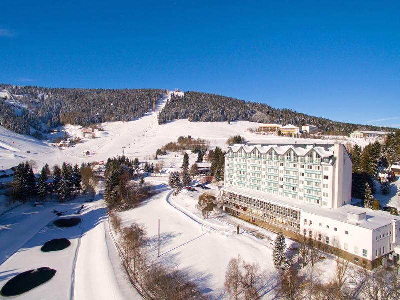 Large hotel beside snow-covered slopes and ski trails under clear blue sky.
