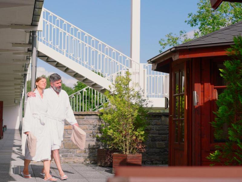 Two people in white bathrobes walk in the outdoor area of a hotel in sunshine.