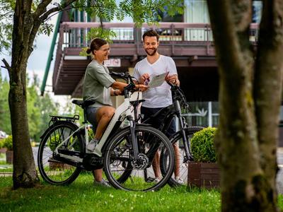 Een stel met e-bikes in een park voor een modern gebouw, omgeven door bomen.