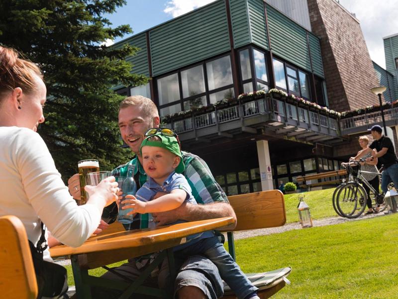 Familie sitzt im Freien an einem Tisch vor einem Hotel, umgeben von Grün und Fahrradfahrern.