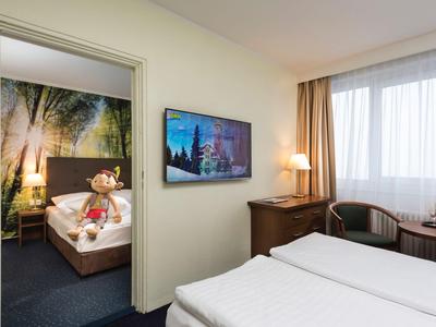 Modern eingerichtetes Hotelzimmer mit einem Doppelbett, einem Fernseher und einem angrenzenden Raum mit Einzelbett.