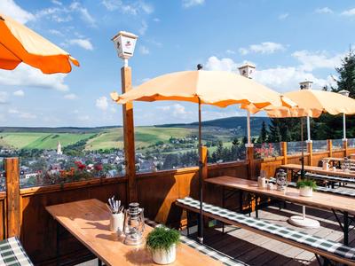 Terrasse mit Holzmöbeln und orangefarbenen Sonnenschirmen mit Blick auf eine Stadt und Hügel im Hintergrund.