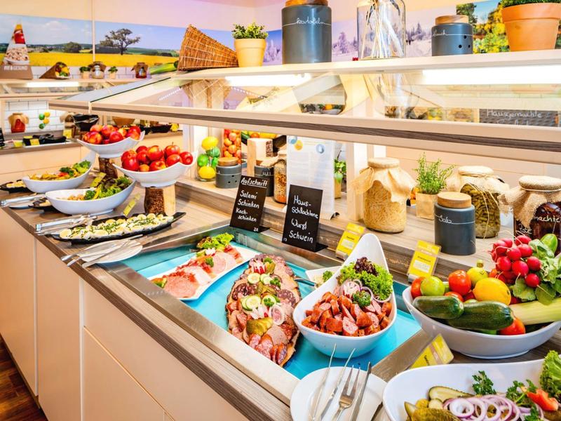 Buffet with fresh fruits, vegetables, cold cuts, and salads in a bright dining area.