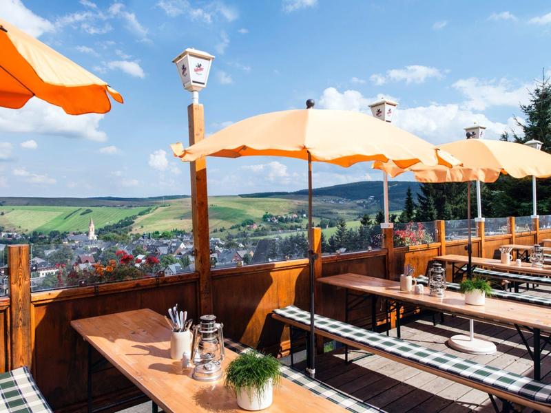 Sunny outdoor terrace with wooden benches and orange umbrellas, overlooking green landscape.