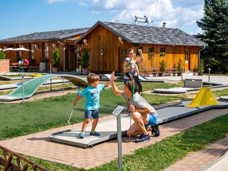 Children playing mini golf on a sunny terrace in front of a wooden building.