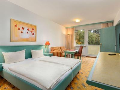 Chambre d'hôtel lumineuse avec deux lits simples, bureau, chaise et balcon donnant sur la verdure.