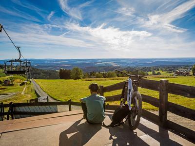 Persoon zit op terras met golfuitrusting en kijkt uit over heuvelachtig landschap onder blauwe lucht.