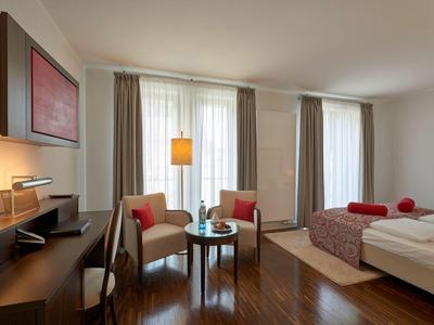 Helles Hotelzimmer mit Bett, zwei Sesseln, Tisch und Schreibtisch mit Lampe.