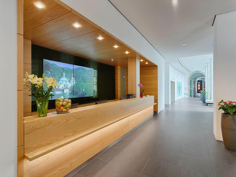 Moderner Hotellobby-Empfang mit Holztheke, Blumen und langen Flur mit Glasfenstern