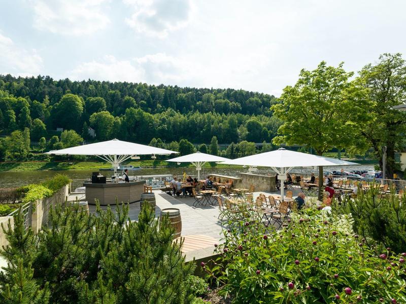 Terrasse mit Sonnenschirmen und Tischen an einem Flussufer, umgeben von Wald und grüner Vegetation.