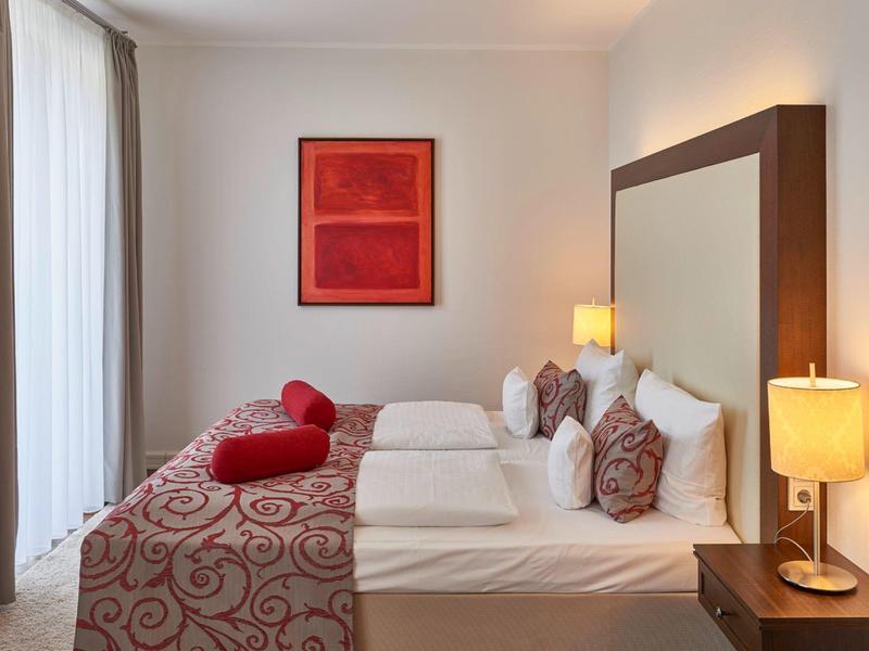 Modern eingerichtetes Hotelzimmer mit Doppelbett, roten Akzenten und zwei Nachttischlampen.