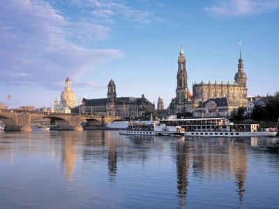 Stadtbild mit historischen Gebäuden und Schiffen auf einem ruhigen Fluss unter blauem Himmel.