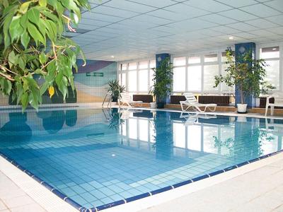 An indoor pool with blue tiles, surrounded by large windows and green plants.