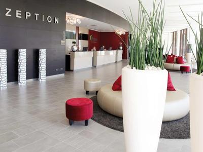 Moderne Hotelrezeption mit grauen Böden, weißer Theke, roten Sitzhockern und großen Pflanzgefäßen.