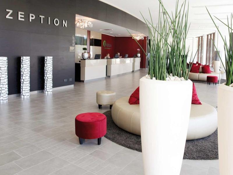 Moderne Hotelrezeption mit grauen Böden, weißer Theke, roten Sitzhockern und großen Pflanzgefäßen.