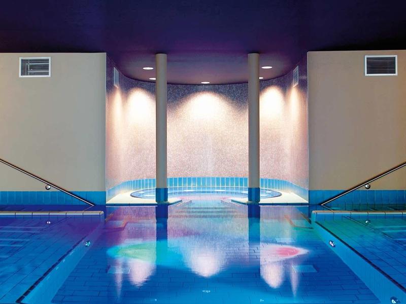 Moderner Innenpool mit Treppen, blauem Wasser, sanfter Beleuchtung und violettem Hintergrund.