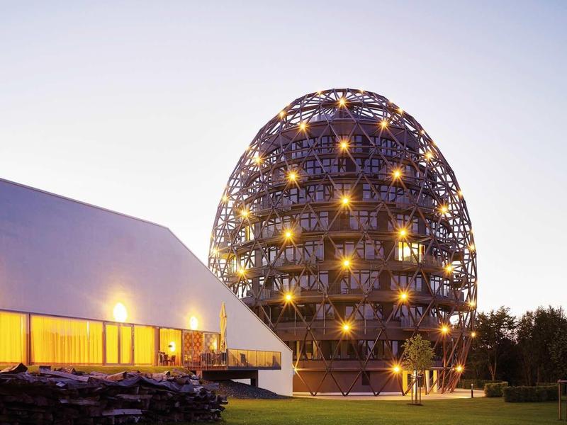 Abendaufnahme: Rundes, beleuchtetes Holzgebäude neben weißem Flachdachbau auf grünem Rasen.